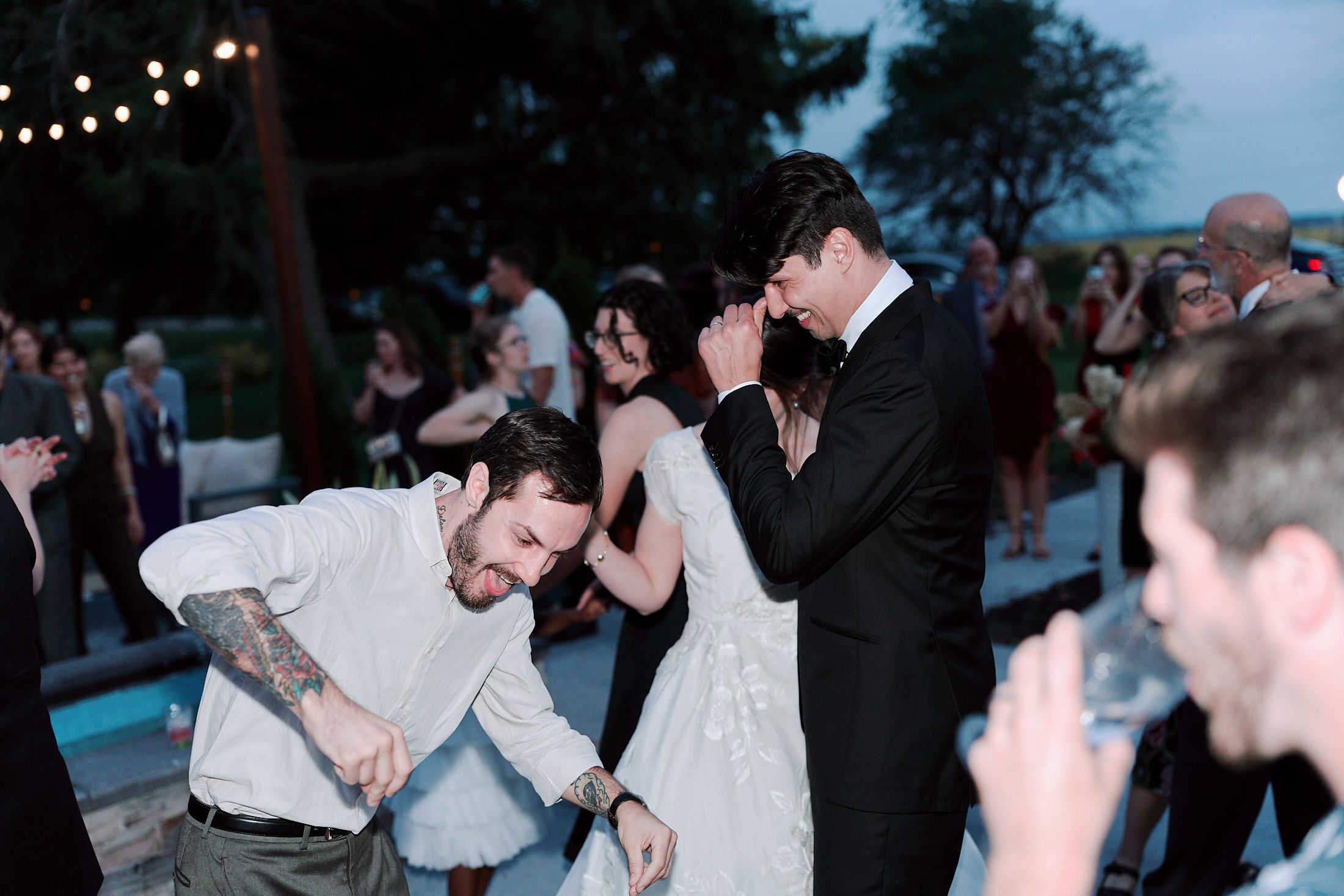 Wedding guests dancing and celebrating at an evening reception under dim lighting.
