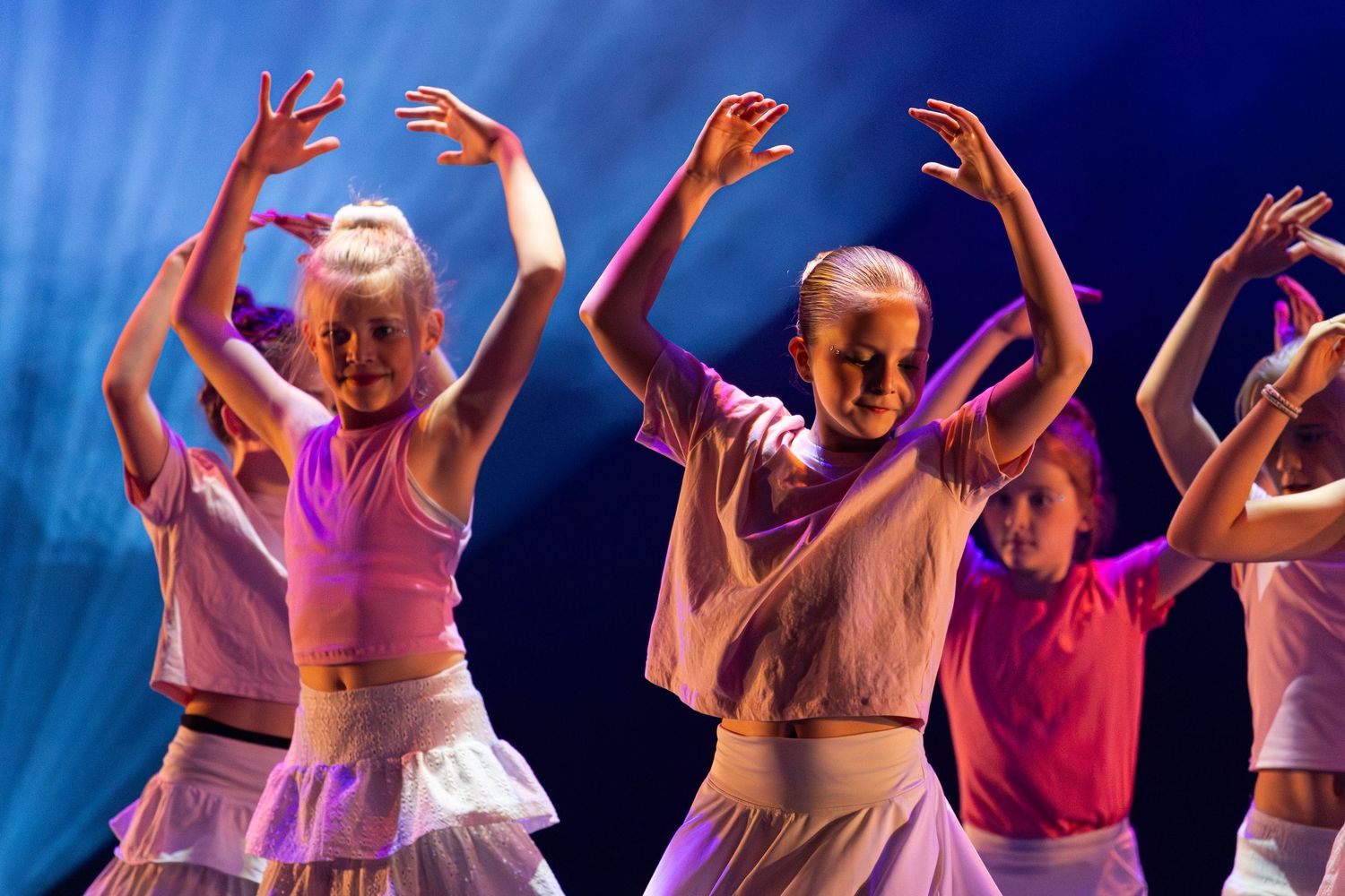 Group of dancers with raised arms perform in unison against a blue-lit backdrop during a stage performance.