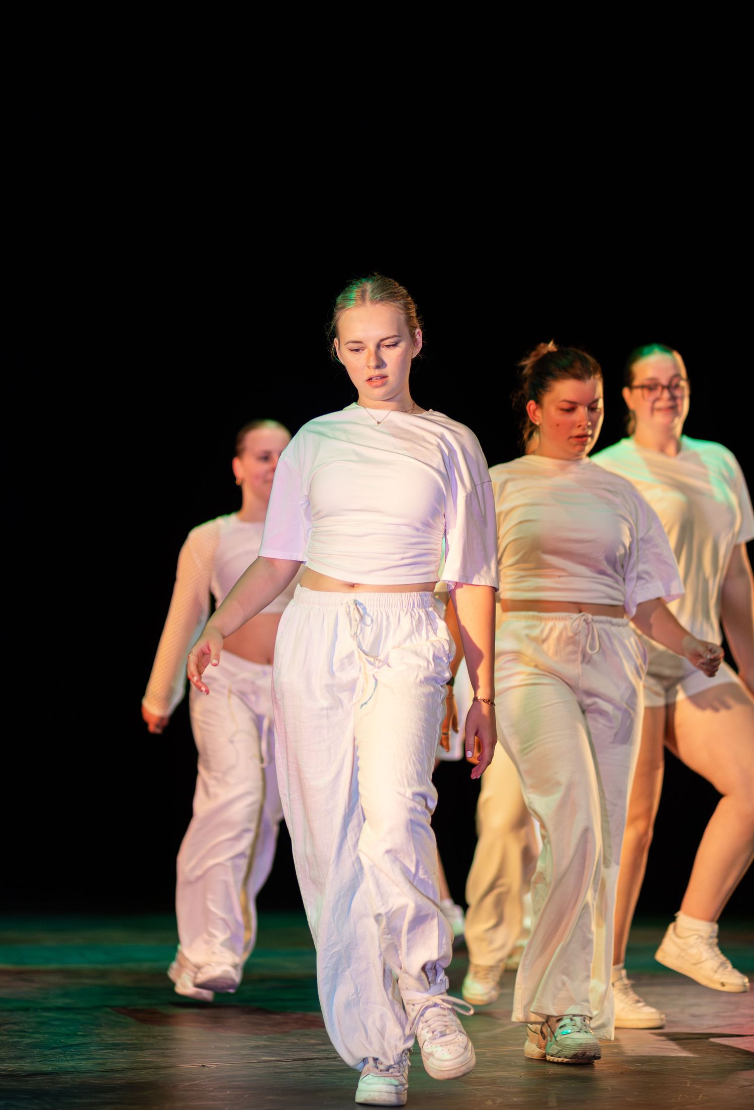 Dancers wearing white outfits perform a synchronized routine on a dark stage with dramatic lighting.