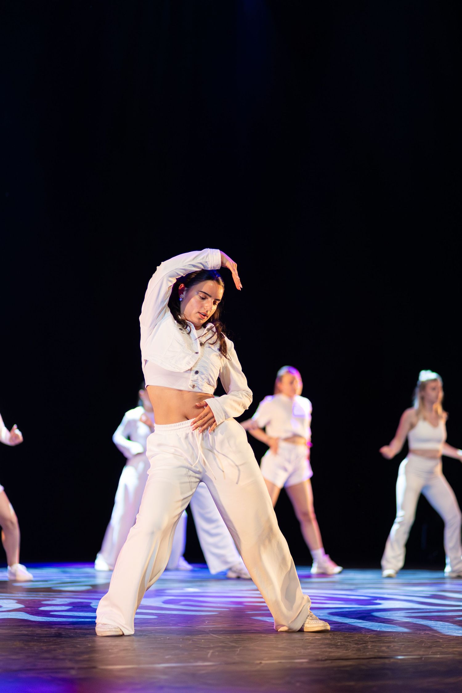 Dancers in white outfits perform dynamic floor movements on an illuminated stage during a modern dance piece.