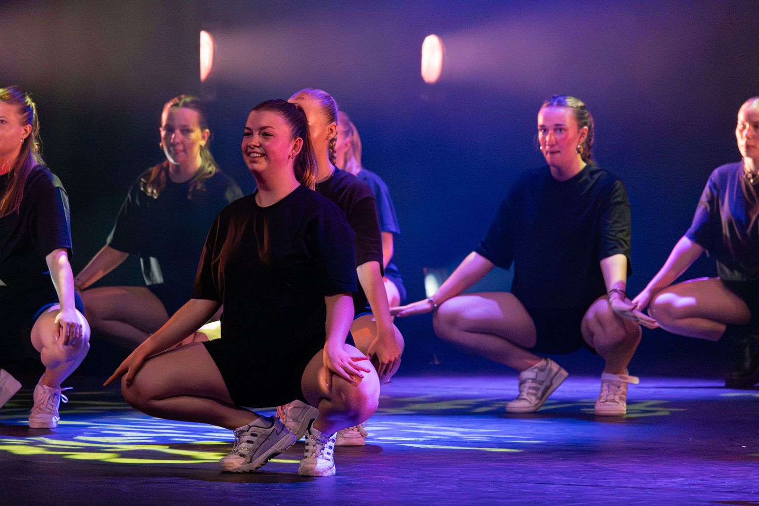 Dancers' feet in black shoes sit on a purple-lit stage during a performance.