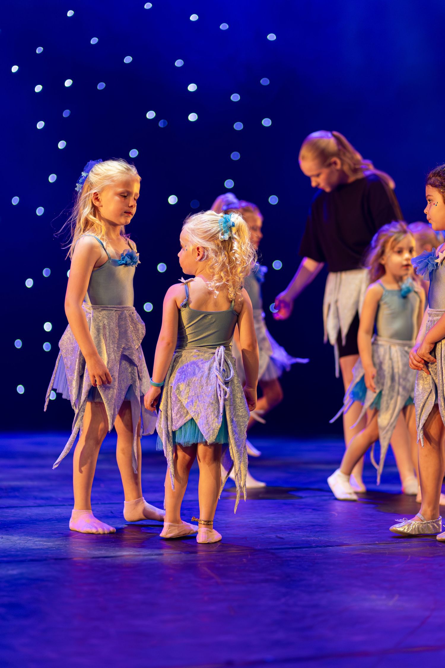 Young dancers in blue dresses perform a synchronized floor routine on a dark stage with twinkling lights in the background.