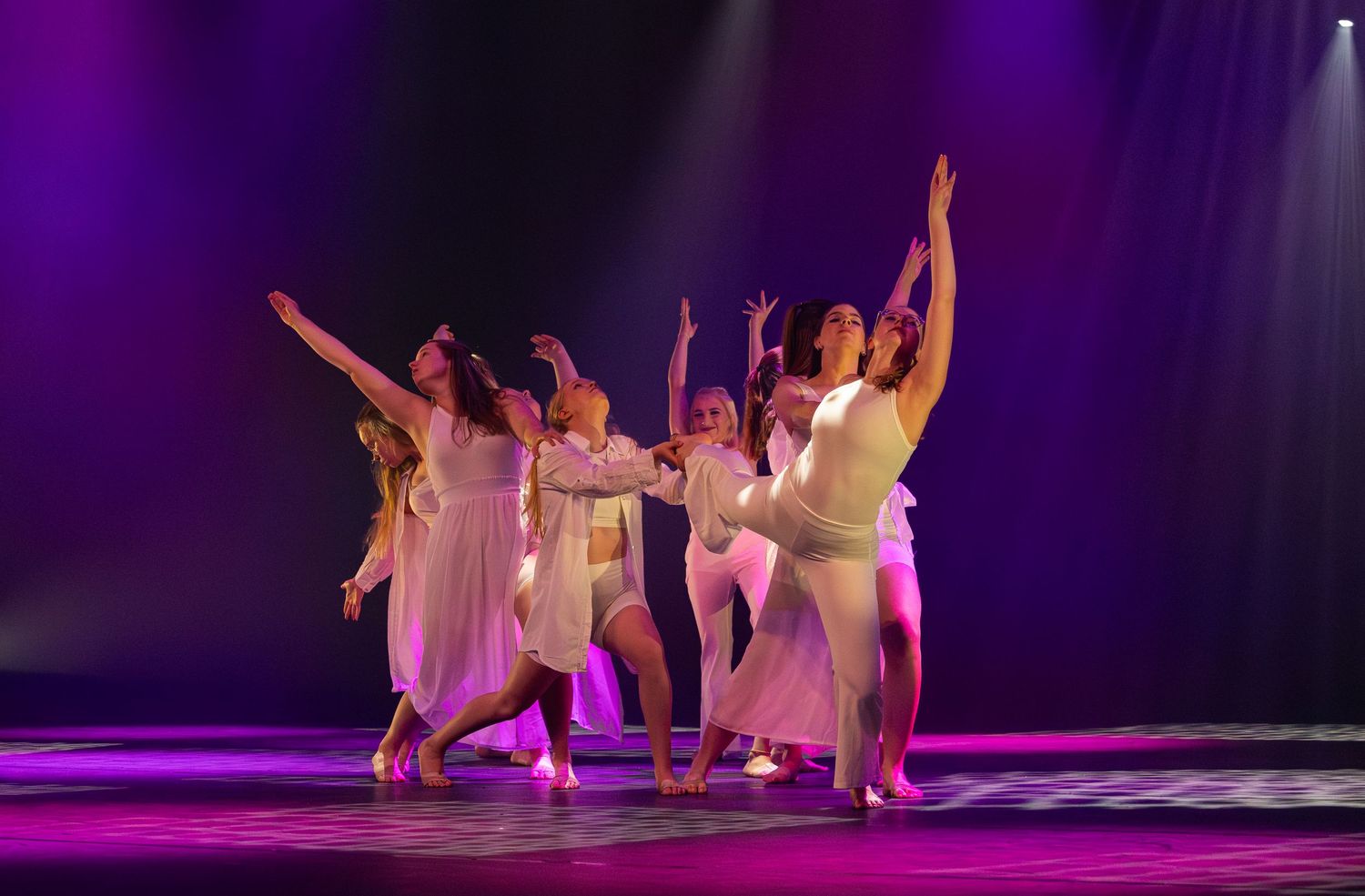 Dancers in white dresses perform on a purple-lit stage during a contemporary dance performance.