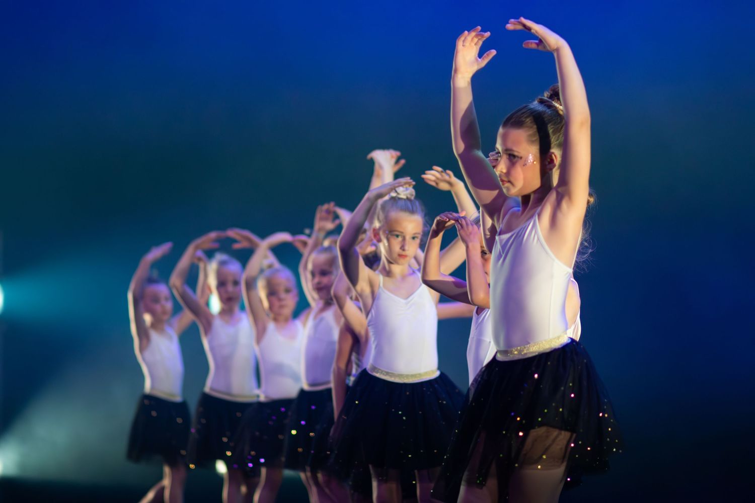 Dancers in white outfits perform a synchronized routine on stage with dramatic blue lighting and atmospheric effects.