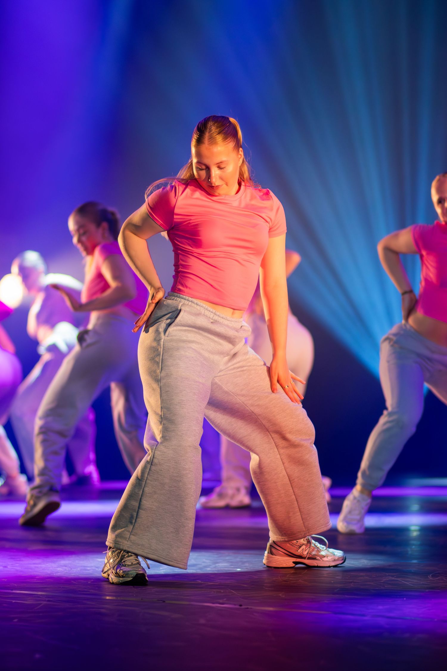 Dancers in pink shirts and gray pants perform low crouching movements on a stage with dramatic blue lighting.