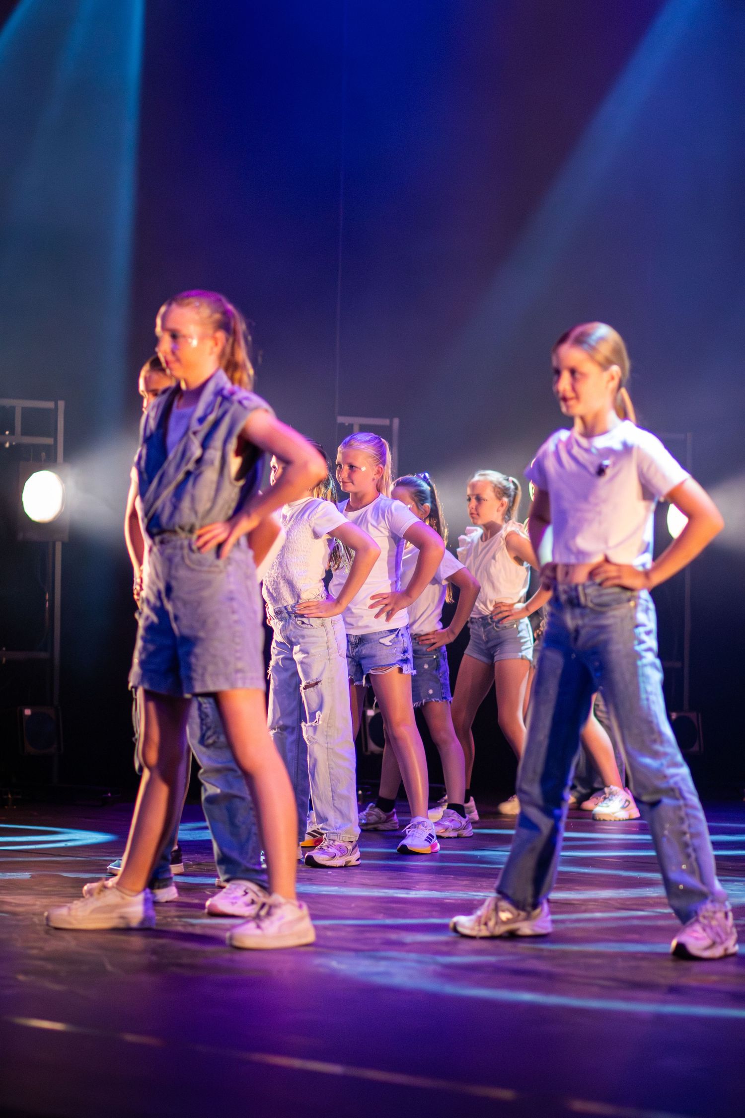 Dancers in casual outfits perform synchronized moves on a purple-lit stage during a hip hop dance routine.