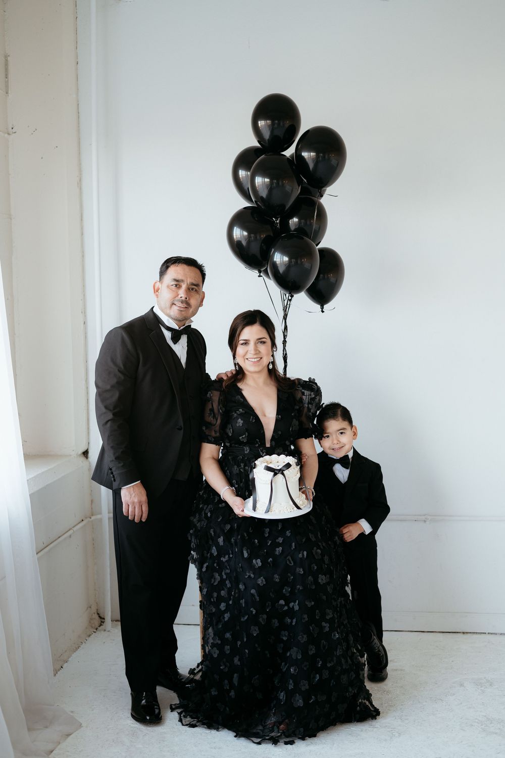 Family portrait with black balloons and formal attire against white backdrop.