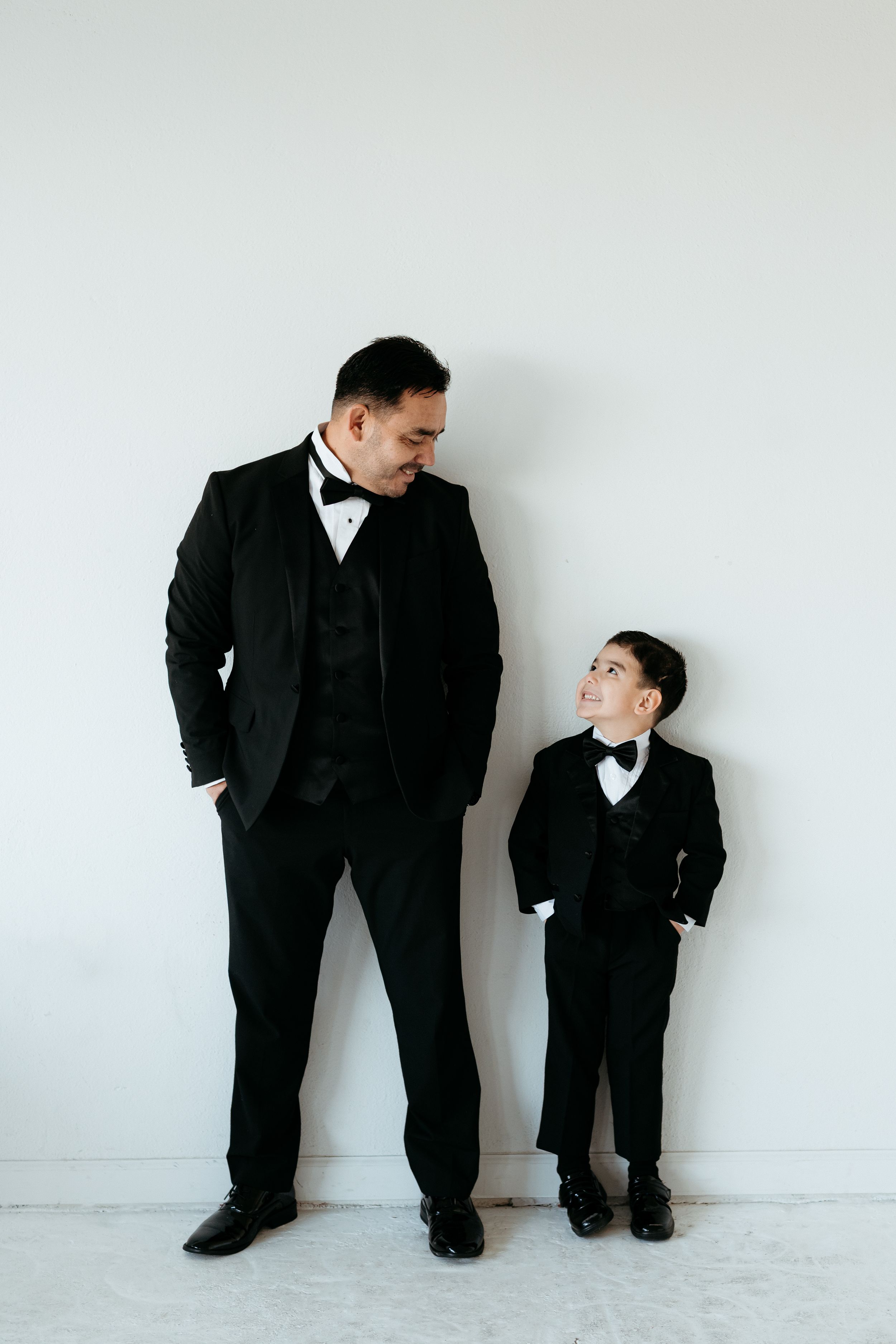 Father and son in black suits interact playfully against white wall.