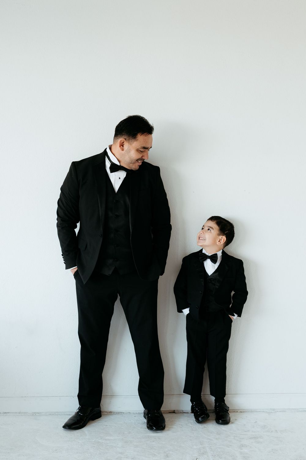 Father and son in black suits interact playfully against white wall.