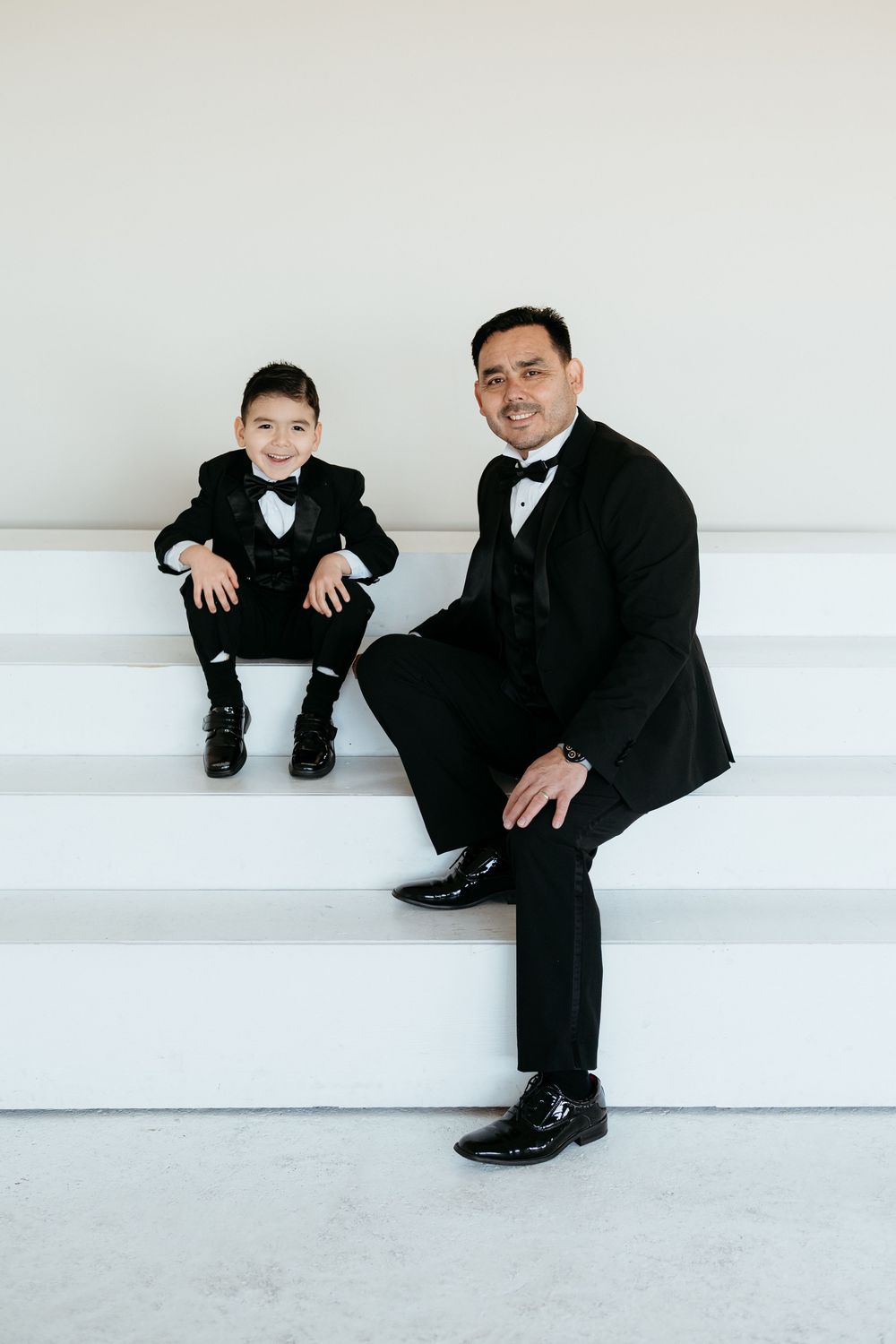 Father and young son in matching tuxedos sit together on white steps.