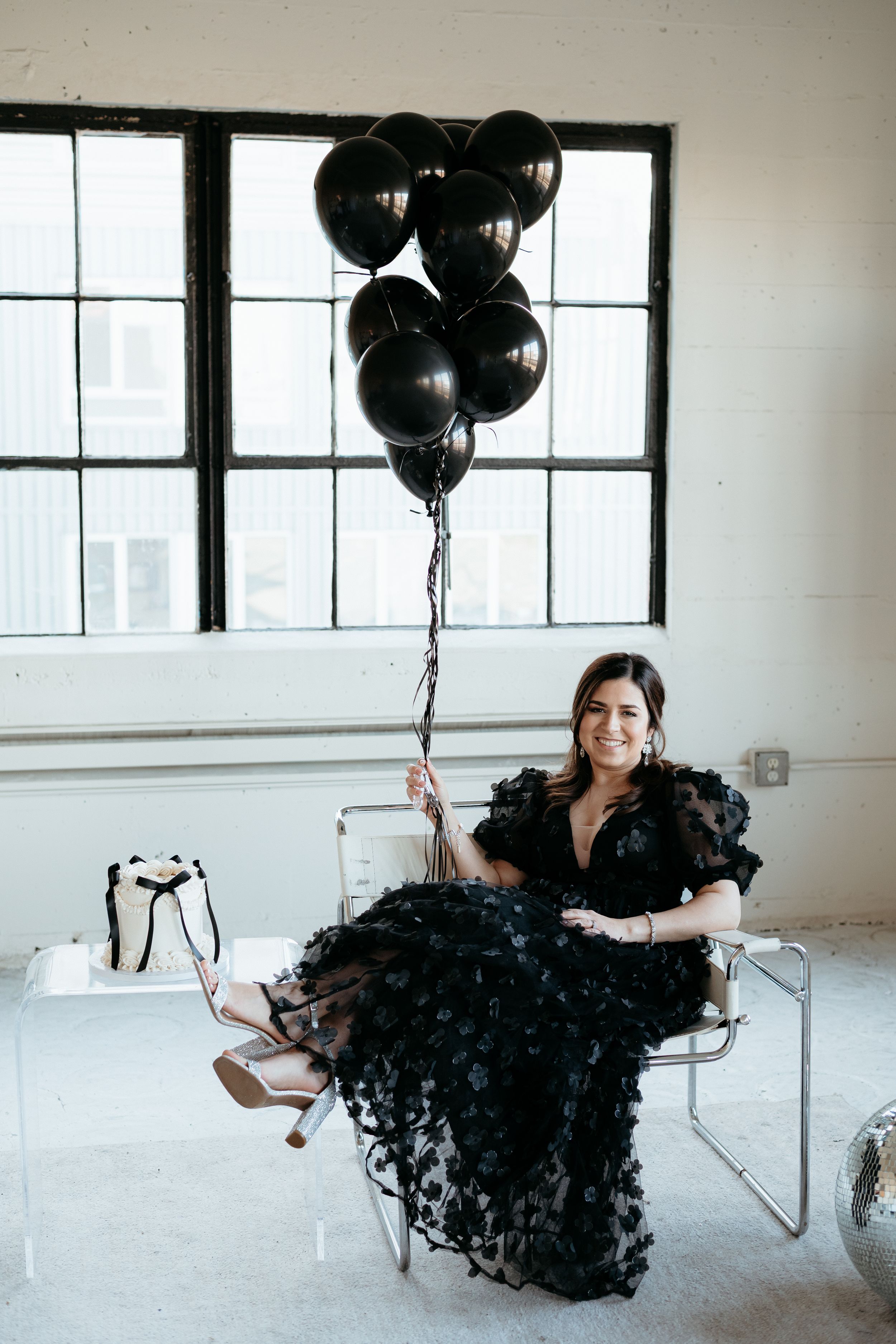 Woman in black textured gown poses with black balloons in minimalist white studio.