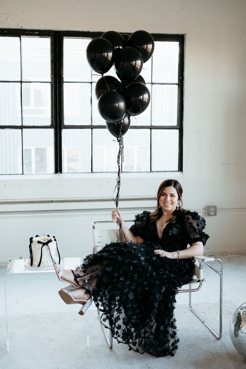 Woman in black textured gown poses with black balloons in minimalist white studio.