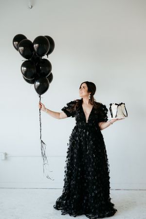 Woman in flowing black dress poses with black balloons in bright studio space.