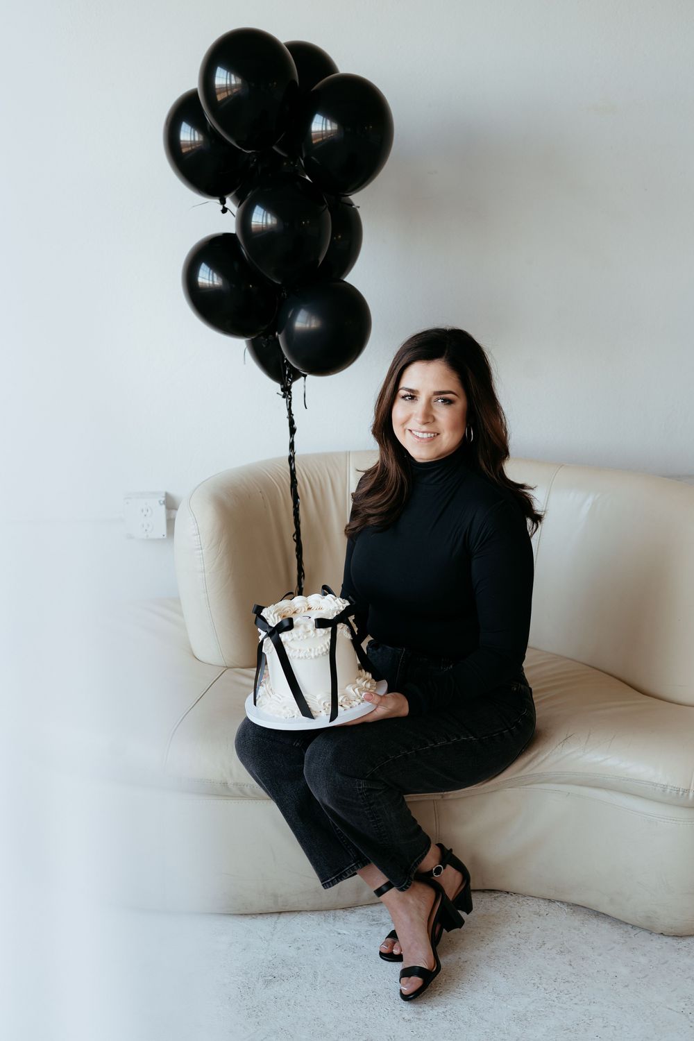 Person in black outfit sits on white couch with black balloons and small cake for a celebration.