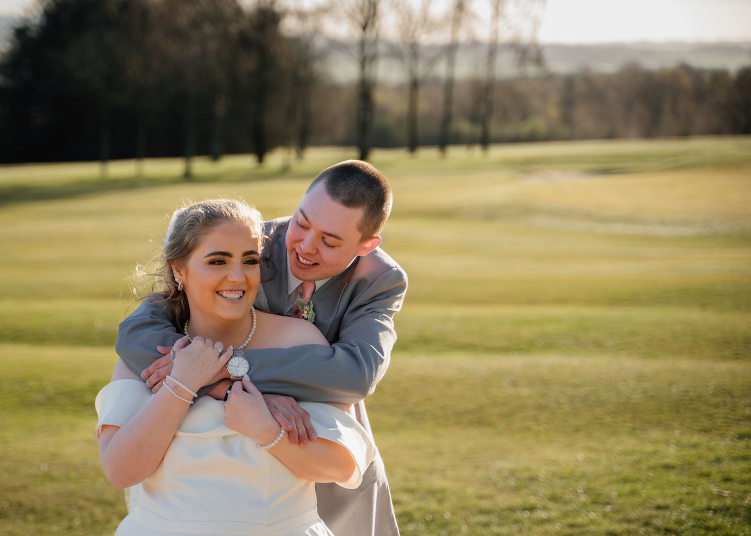 Playful wedding couple shares joyful moment in sunny meadow against green landscape.
