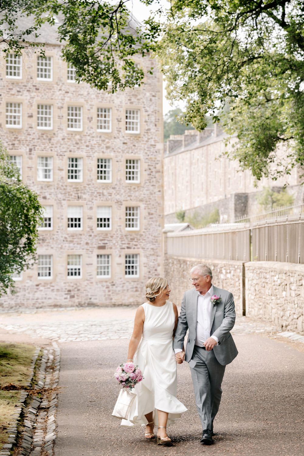 Playful wedding couple shares joyful moment in sunny meadow against green landscape.
