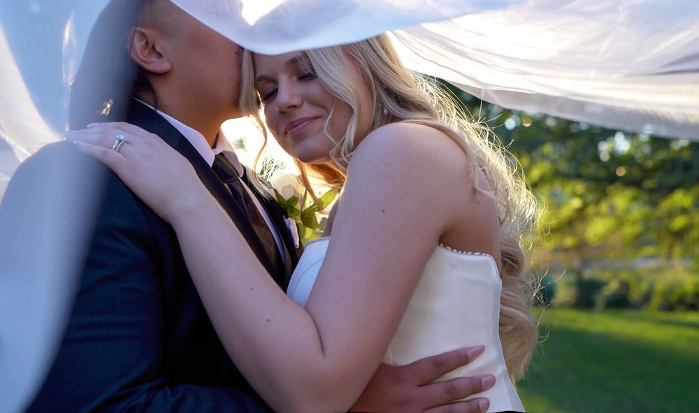 A romantic wedding couple embraces outdoors during golden hour with sunlight streaming through a sheer canopy.