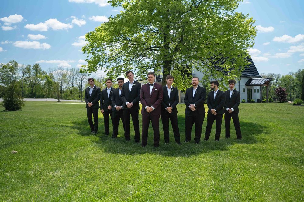 Groom and groomsmen in black and burgundy suits posing outdoors on green lawn with large tree and white barn.
