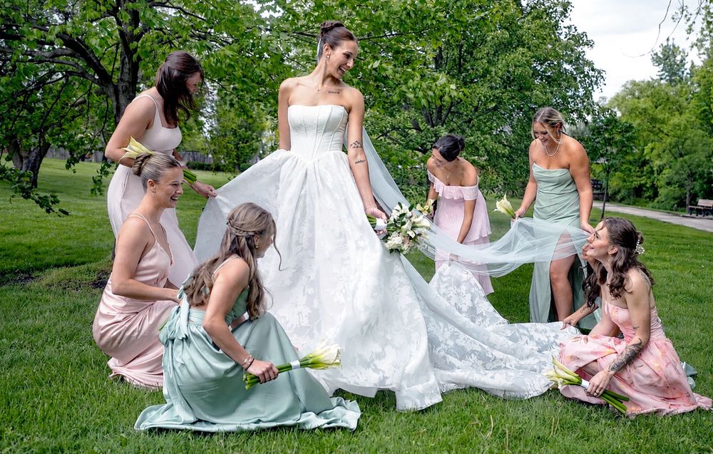 Bride in white gown surrounded by bridesmaids in pink and mint dresses arranging her train on green grass outdoors.