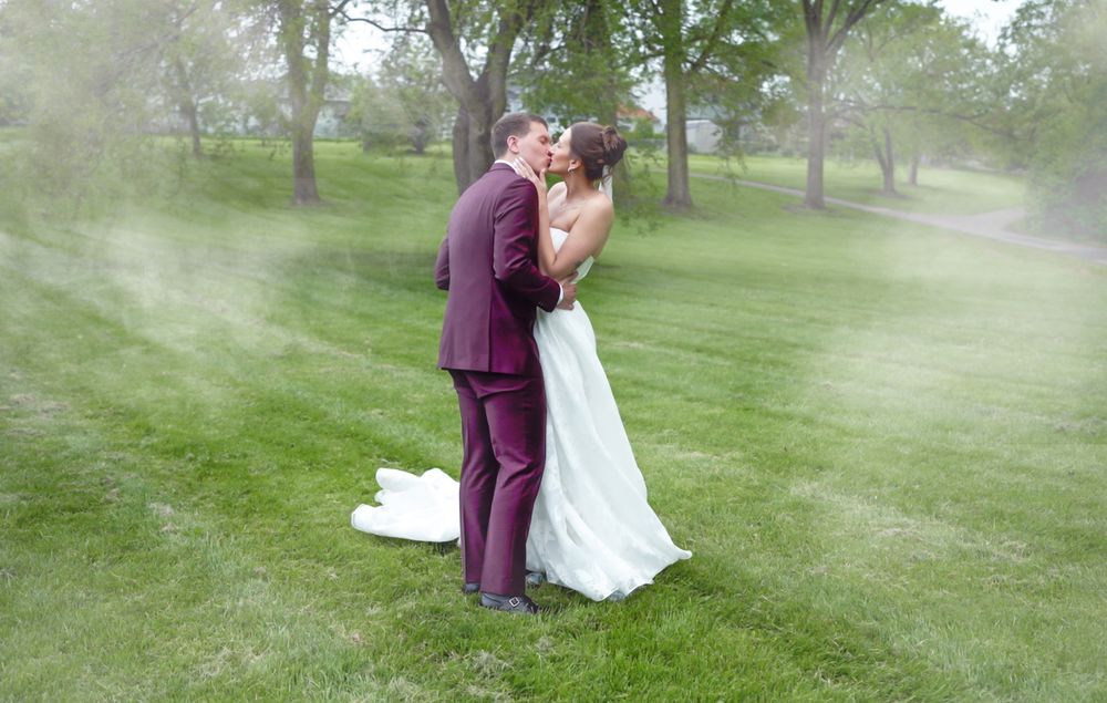 Bride and groom share a romantic kiss on lush green grass, surrounded by a dreamy mist, with trees in the background.