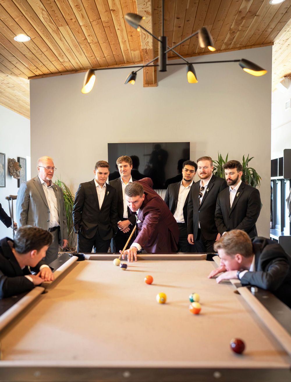 Groomsmen in black tuxedos gather around a pool table watching the groom in burgundy jacket take a shot.