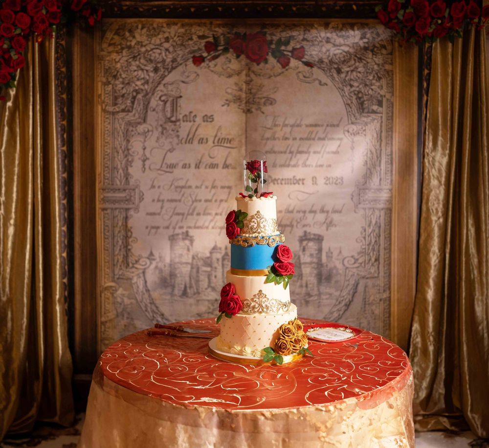 Elegant multi-tier Beauty and the Beast themed wedding cake with red roses on an ornate gold table.