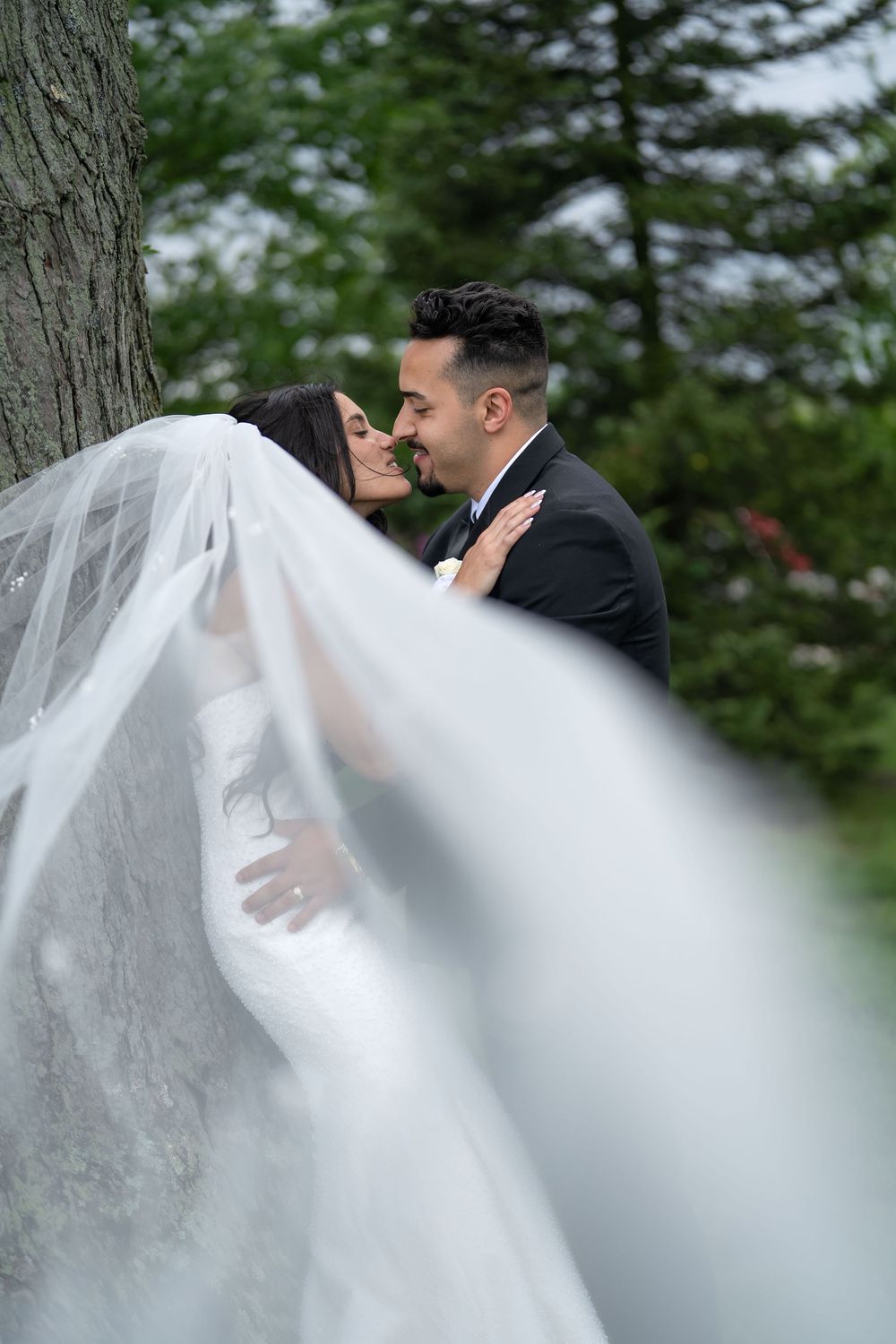 Bride and groom share a romantic moment near a tree, her flowing veil billowing in the foreground.