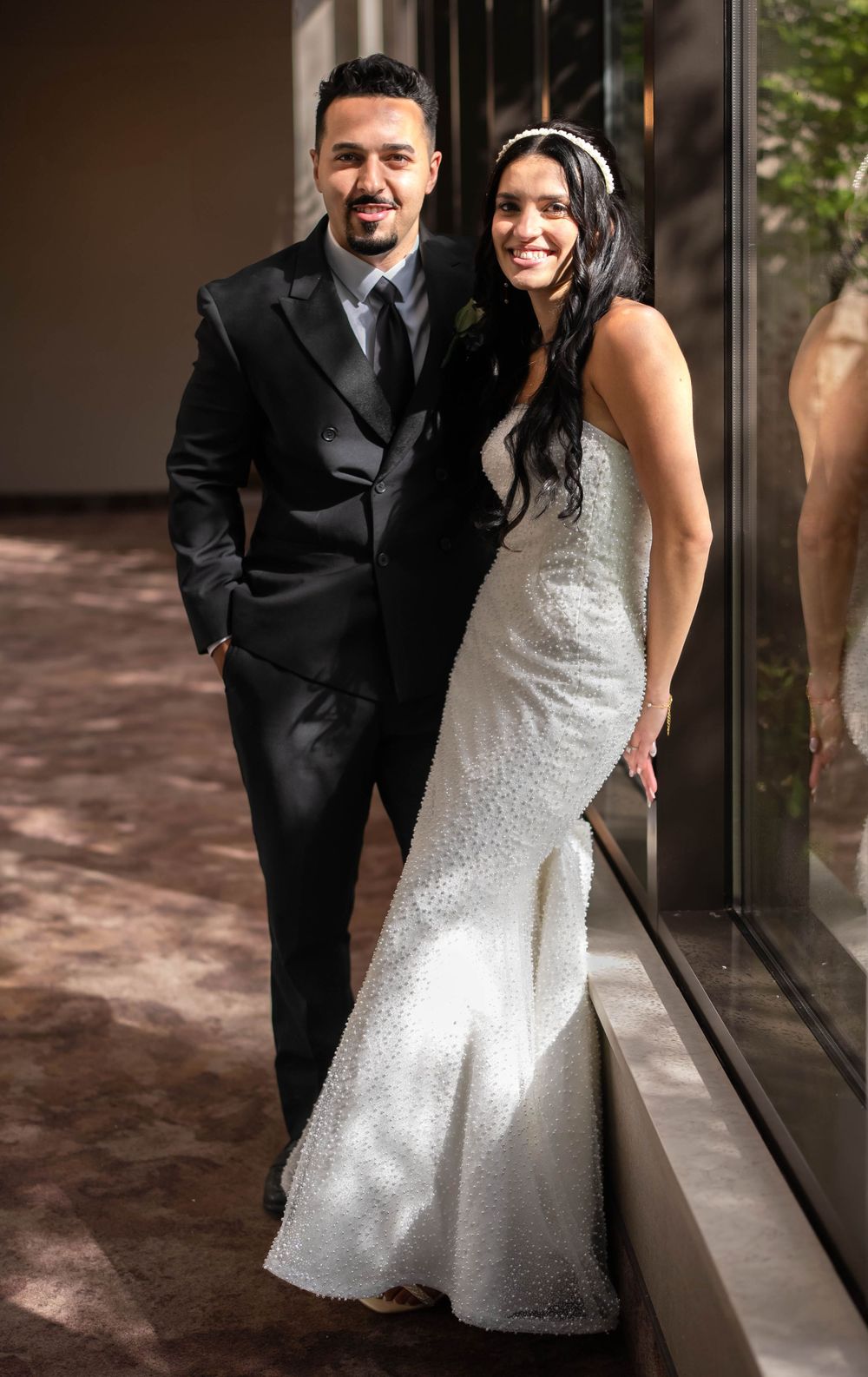 A smiling couple poses in wedding attire, groom in black tuxedo and bride in white beaded gown with headpiece.
