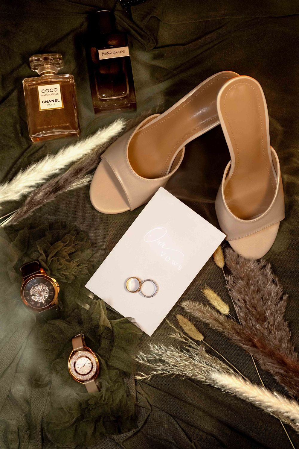 Elegant wedding flat lay with Chanel perfume, nude heels, gold rings, watches, and pampas grass on green silk.