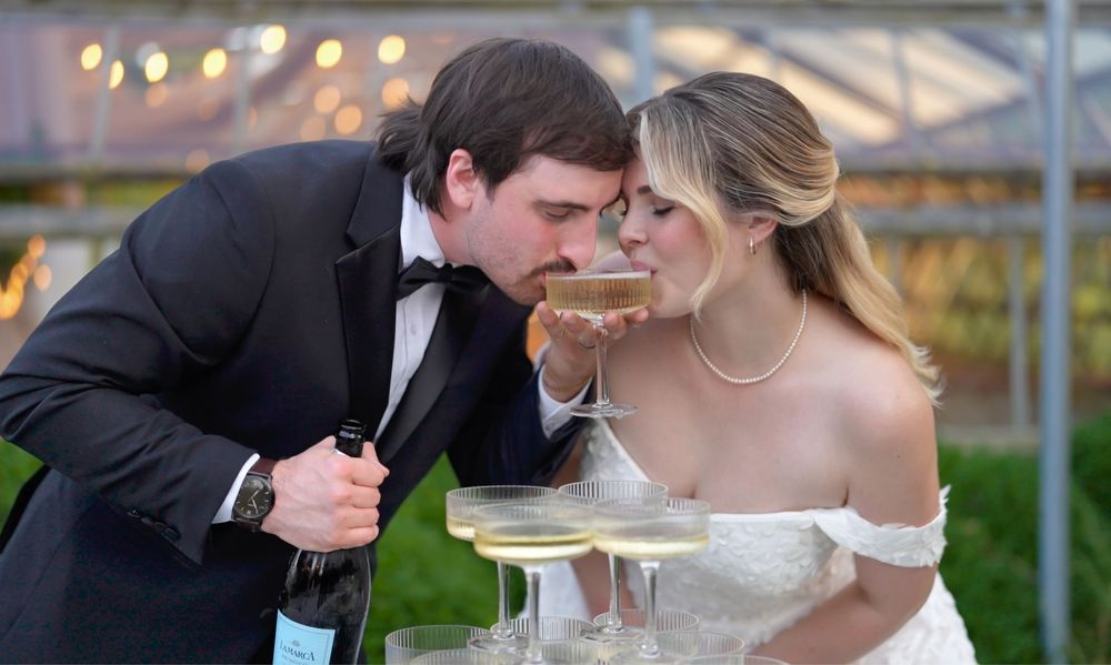 Newlyweds share a playful moment while drinking champagne at their outdoor wedding reception.