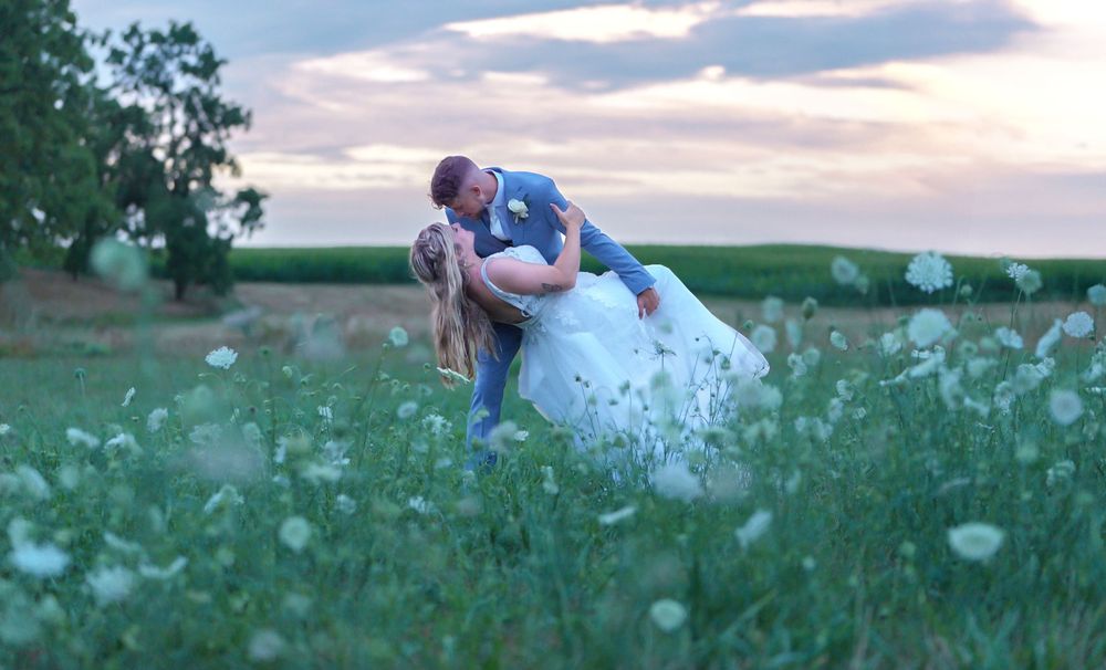 Groom dipping bride in a romantic kiss among white wildflowers in a lush green field at sunset.