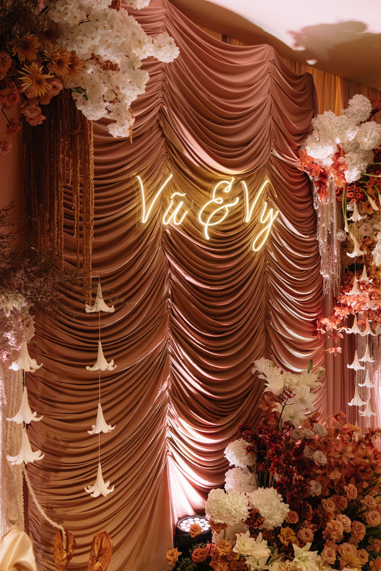 Elegant rose-gold draped wedding backdrop with neon sign reading Vu and Vy surrounded by flowers.