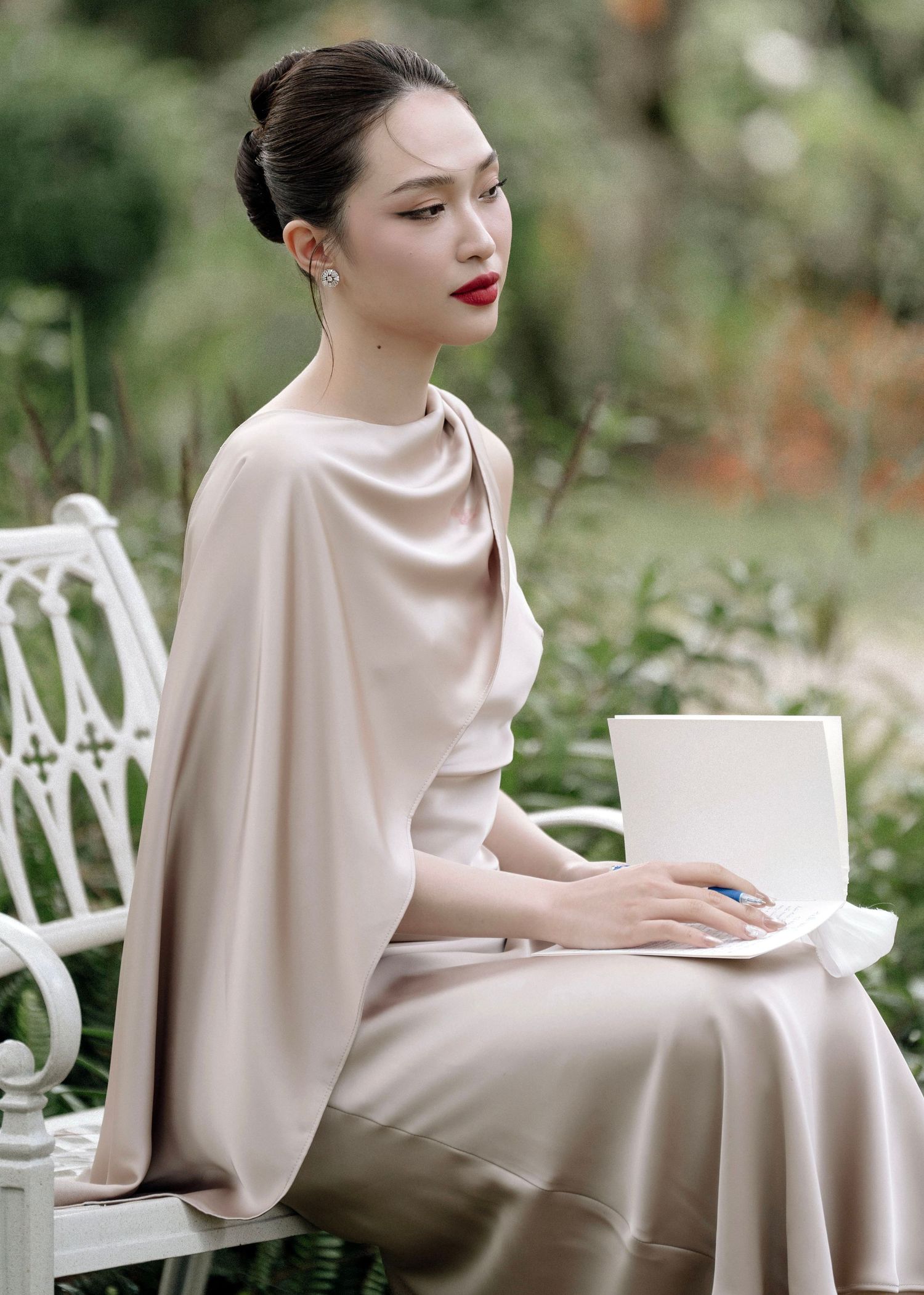 Elegant woman in champagne satin cape dress seated on white bench in garden.