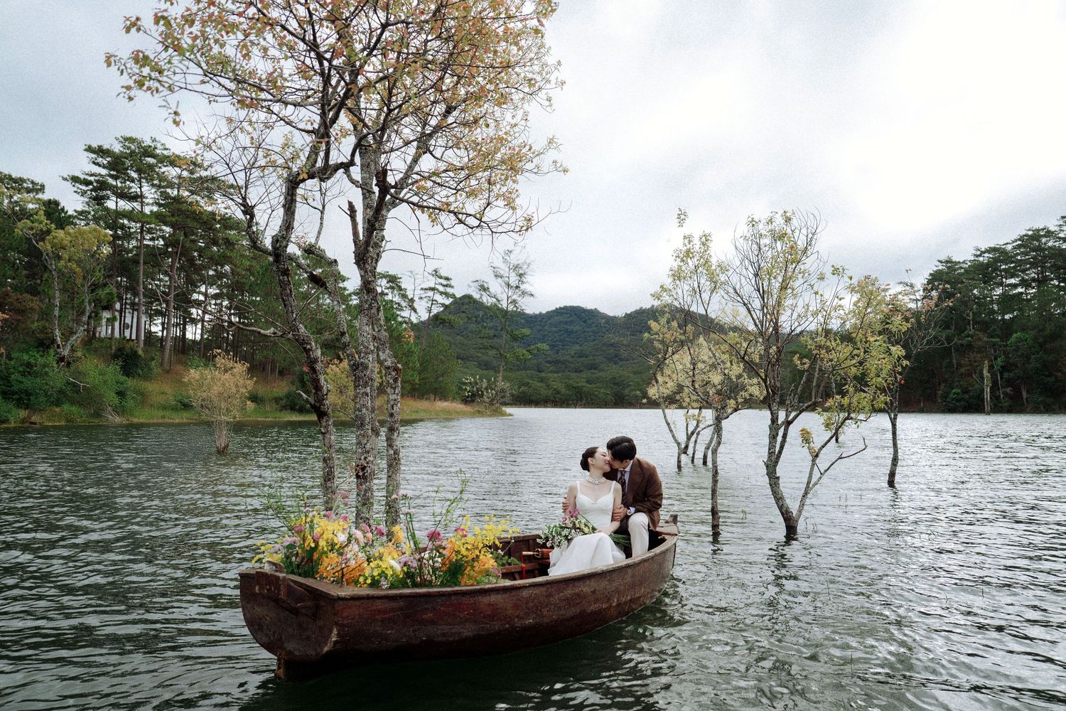 Bride and groom kiss in flower-filled wooden boat on scenic lake with bare trees and mountains behind.