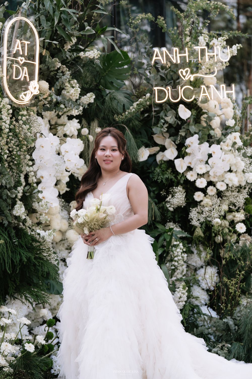 Smiling bride in a white ruffled gown holds a bouquet in front of a floral wedding backdrop.