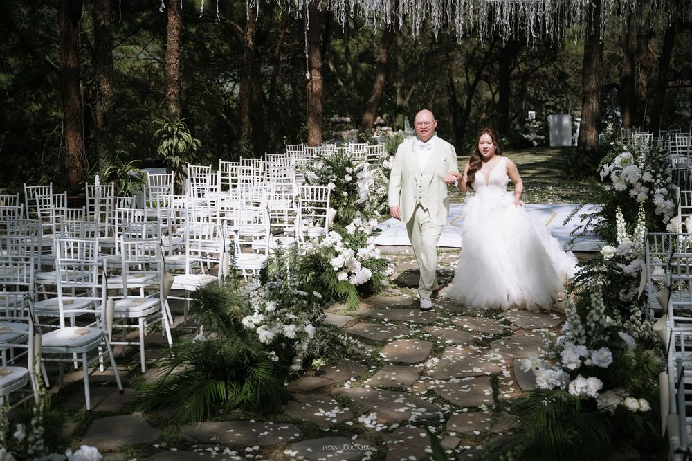 Bride and groom walking down an outdoor forest wedding aisle lined with white chairs and flowers.