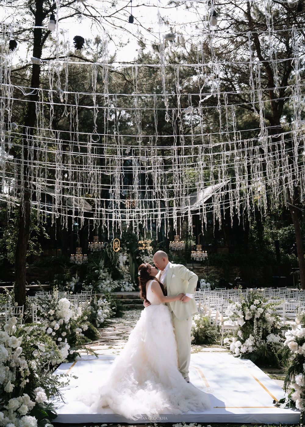 Newlyweds sharing first kiss under cascading white ribbon canopy at enchanting outdoor forest wedding.