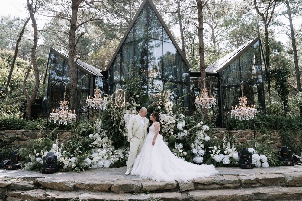 Bride and groom posing before a glass chapel surrounded by lush greenery and white floral arrangements.