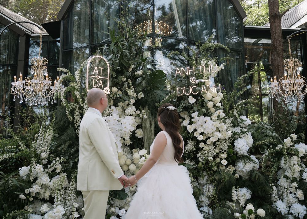 Couple holding hands before elaborate white floral backdrop with golden chandeliers at outdoor wedding.