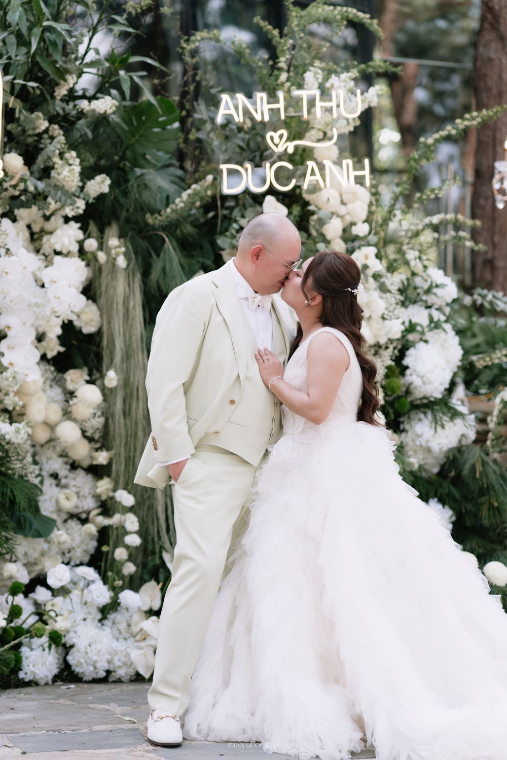 Bride and groom share a kiss in front of a lush white floral backdrop at the Anh Thu and Duc Anh wedding.