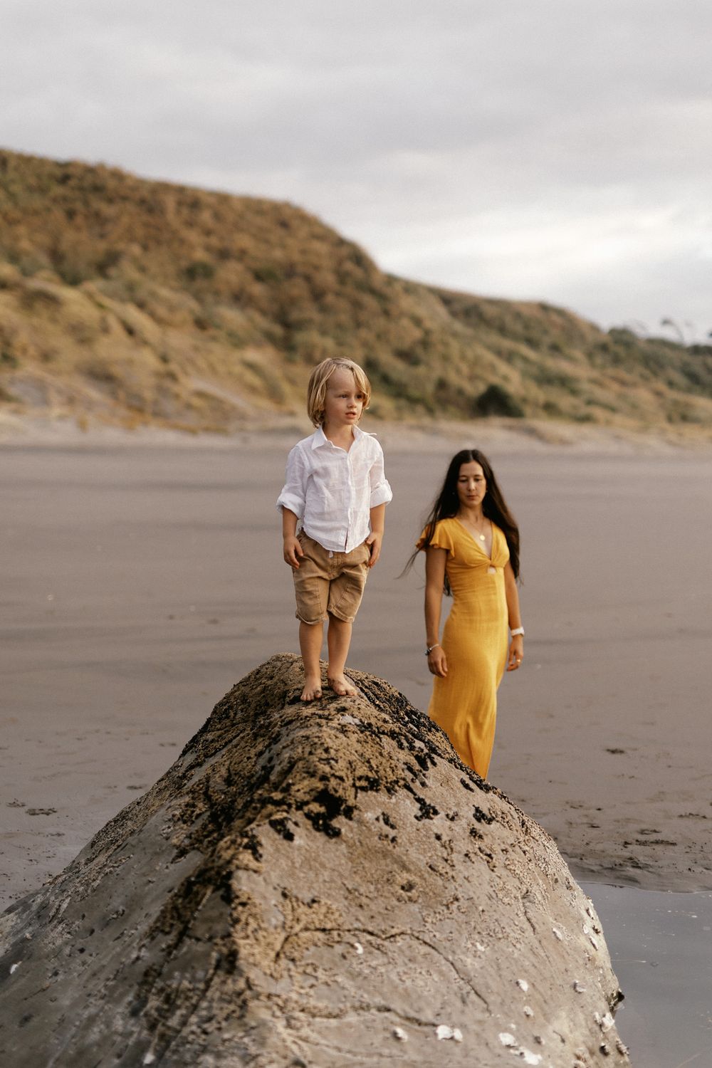 People exploring and standing on large rocks at a coastal beach with hills in the background.
