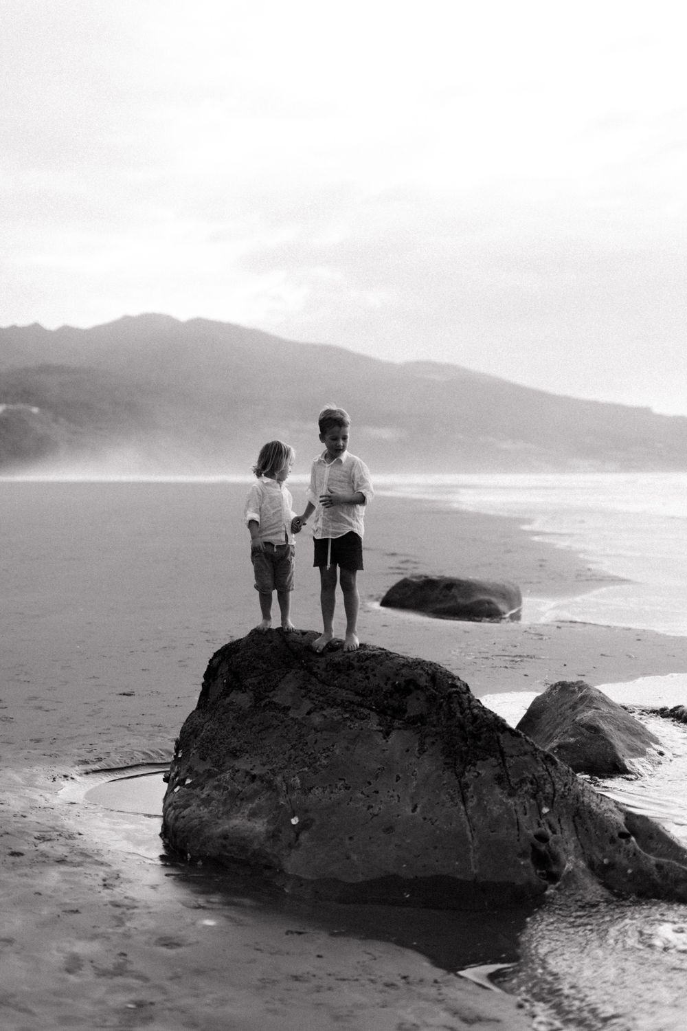 Black and white coastal scene showing people standing on large rocks at the beach with mountains in the distance.
