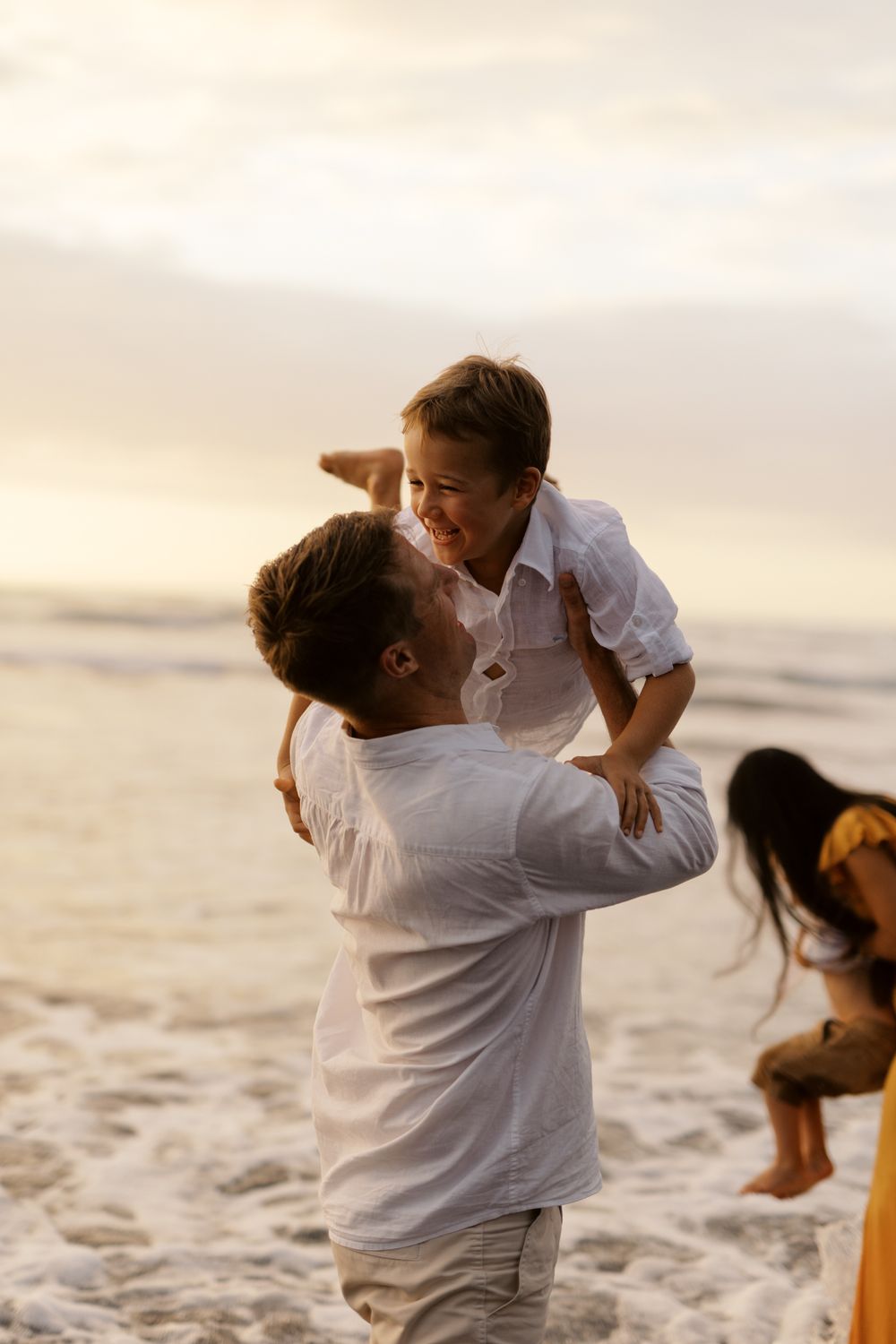 A couple in white embraces tenderly on a sunlit beach with a dog visible in the background.