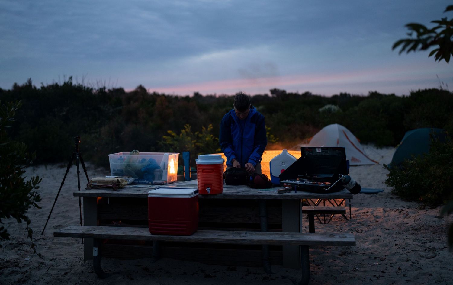 Camping scene at dusk showing tents and campfire glowing under darkening sky.