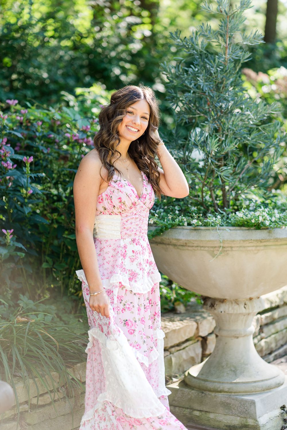 A romantic garden photoshoot with a pink floral dress near stone planters and greenery.