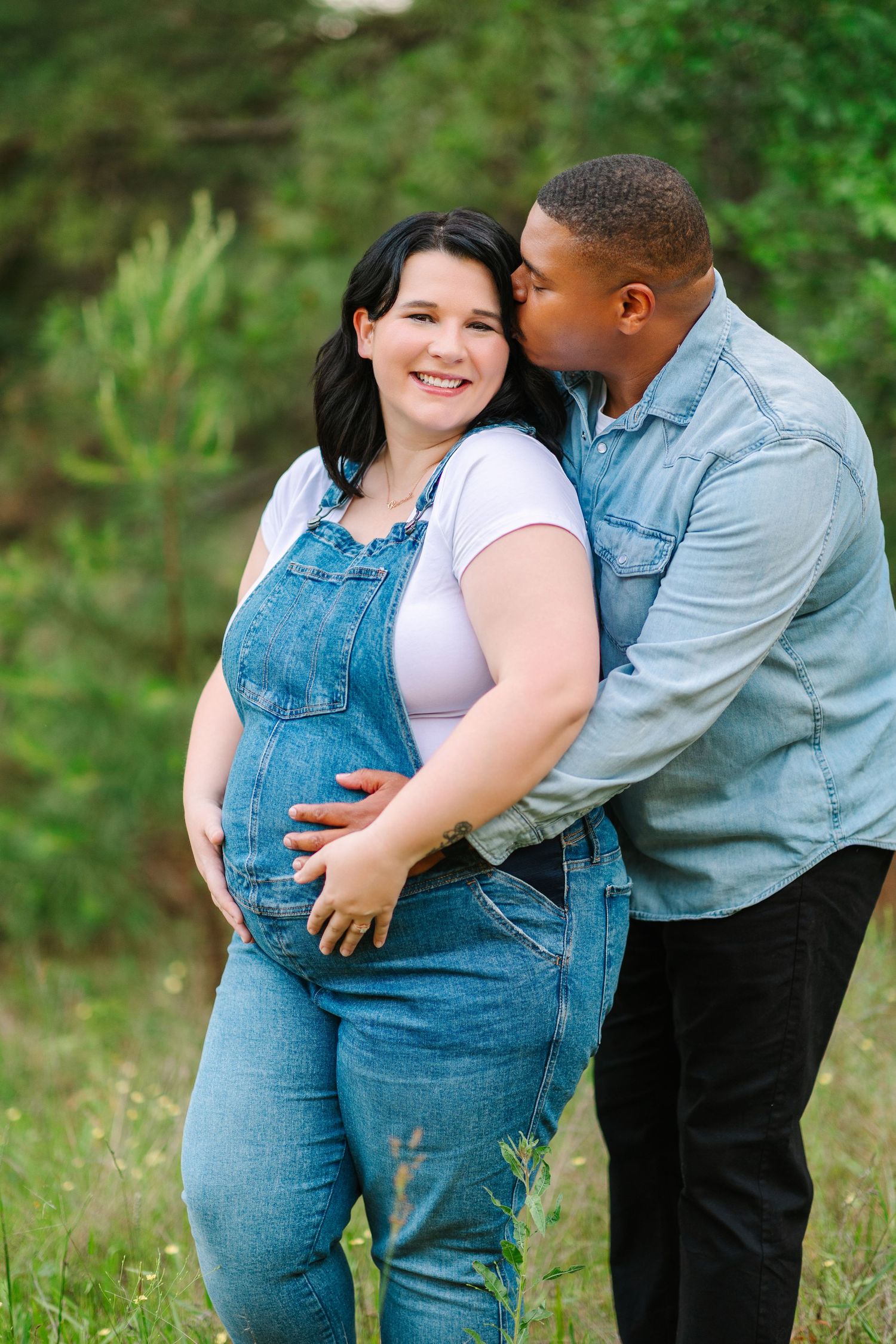 A couple shares tender moments during an outdoor maternity photoshoot in a lush green setting.