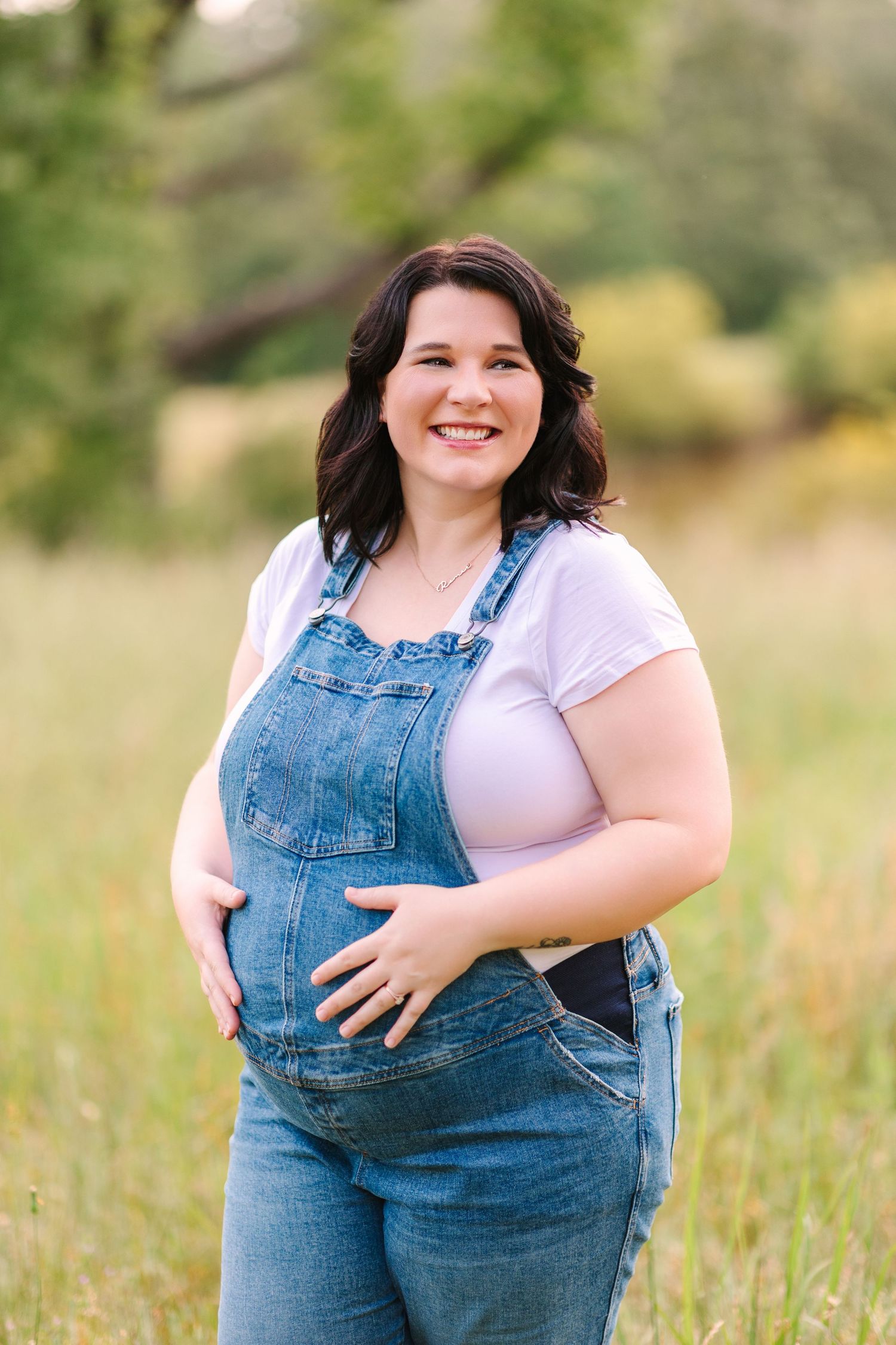 Person wearing denim overalls and white shirt poses joyfully in a grassy outdoor field on a sunny day.