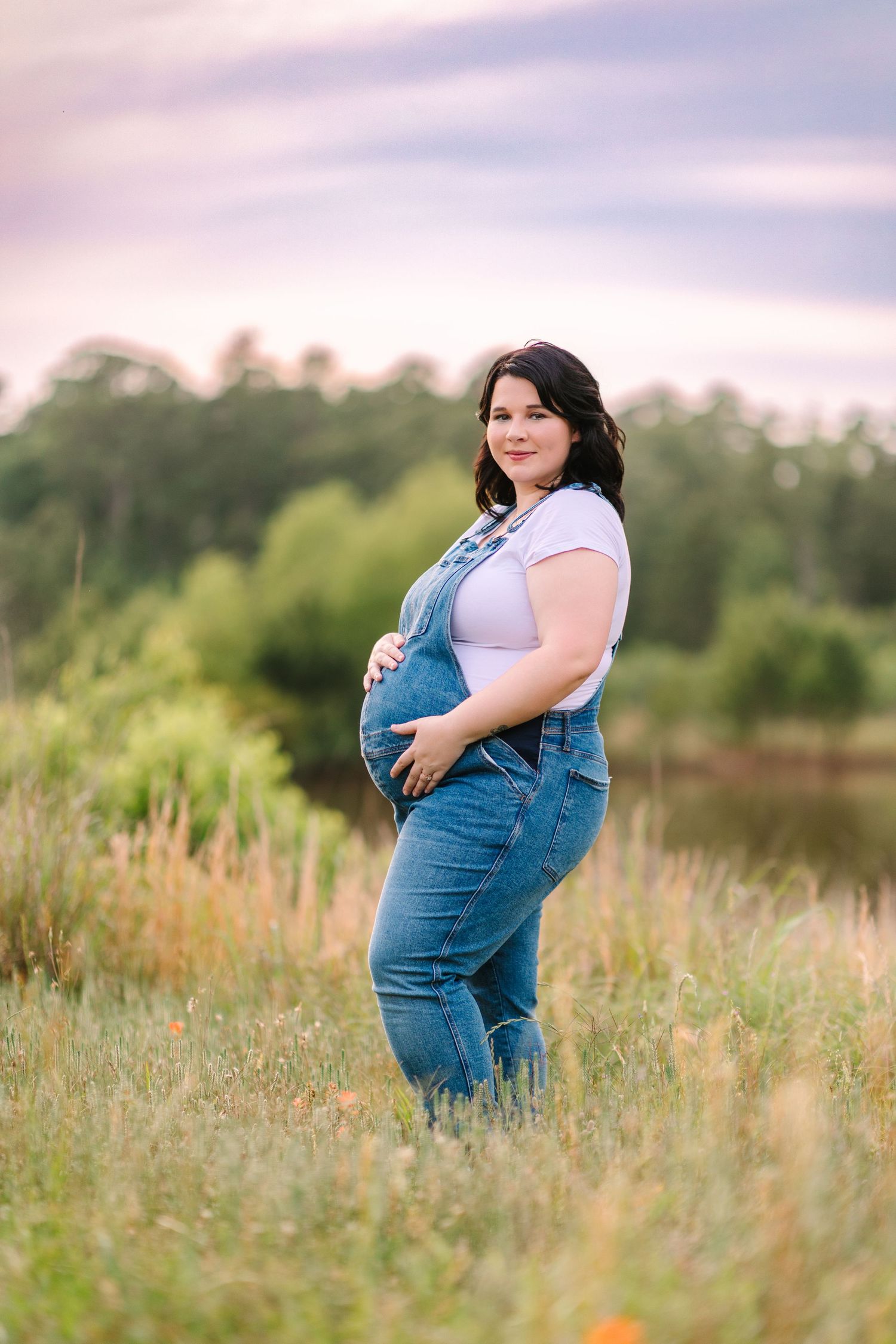 A person in denim overalls poses in a sun-lit meadow during a maternity photography session.