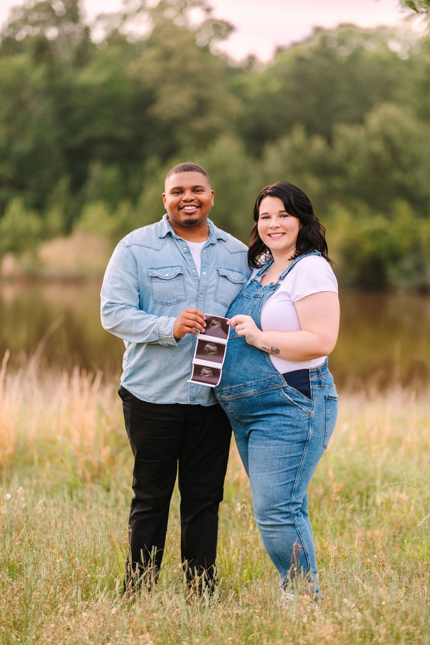 Couple holding an ultrasound photo stands in a grassy field during a pregnancy announcement photoshoot.