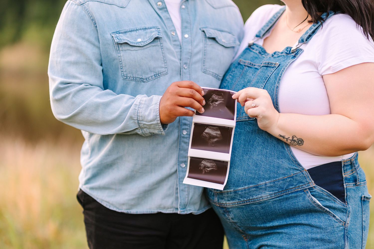 A couple holds an ultrasound photo in a sunlit outdoor field wearing blue denim and pink clothing.